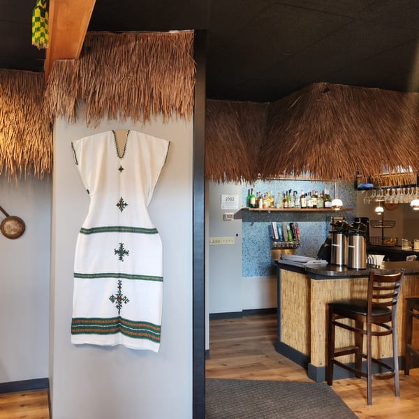 A white Ethiopian dress with green and gold embroidery hangs next to a bar with thatched decor.